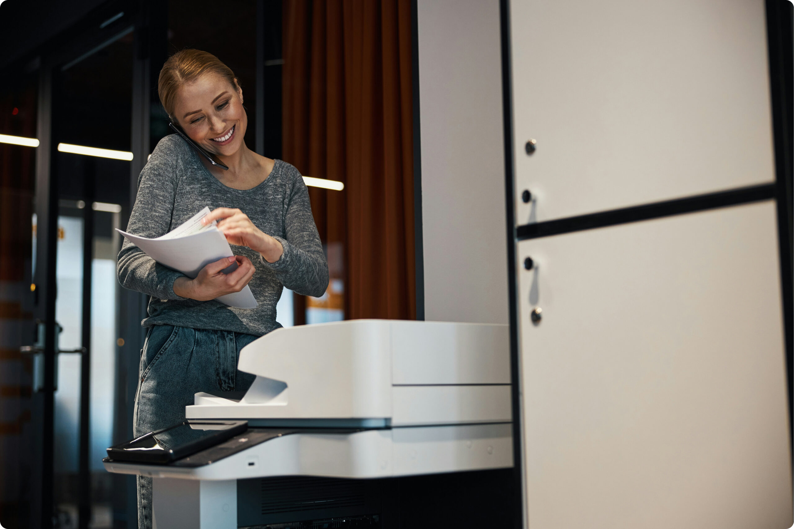 Getty Unsplash — a woman consulting printed documents near a multifunction printer in a modern office environment.
