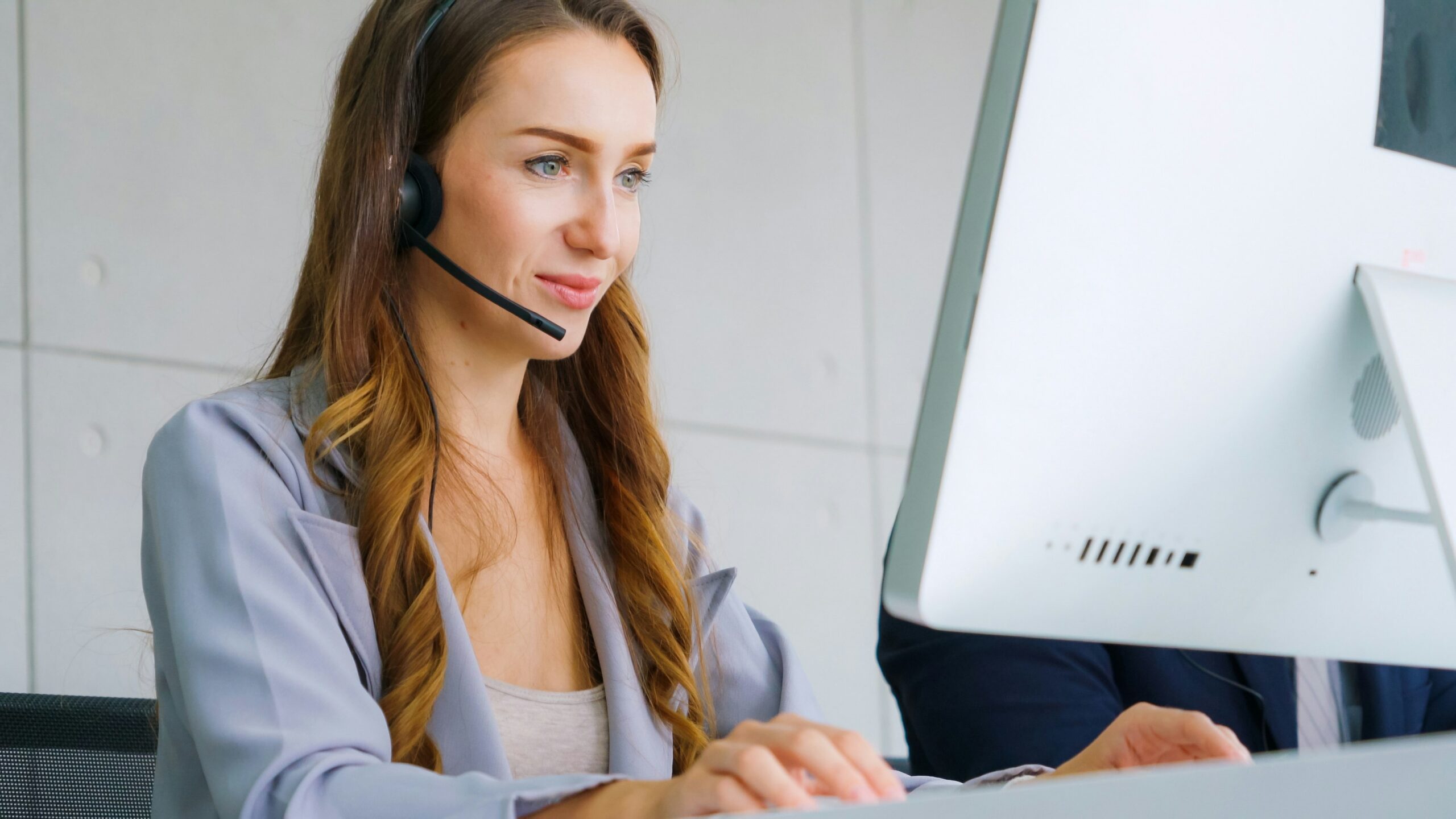 Woman wearing customer service headset in professional attire, working at desktop computer in a business environment.