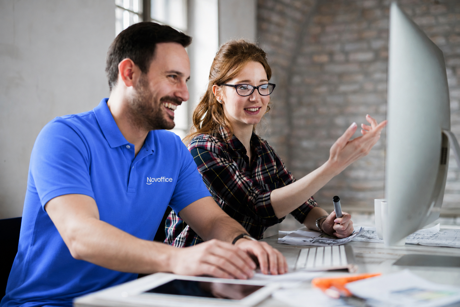 A Novoffice technician in a blue polo shirt helps a customer set up her workstation by showing her computer screen in a modern office.